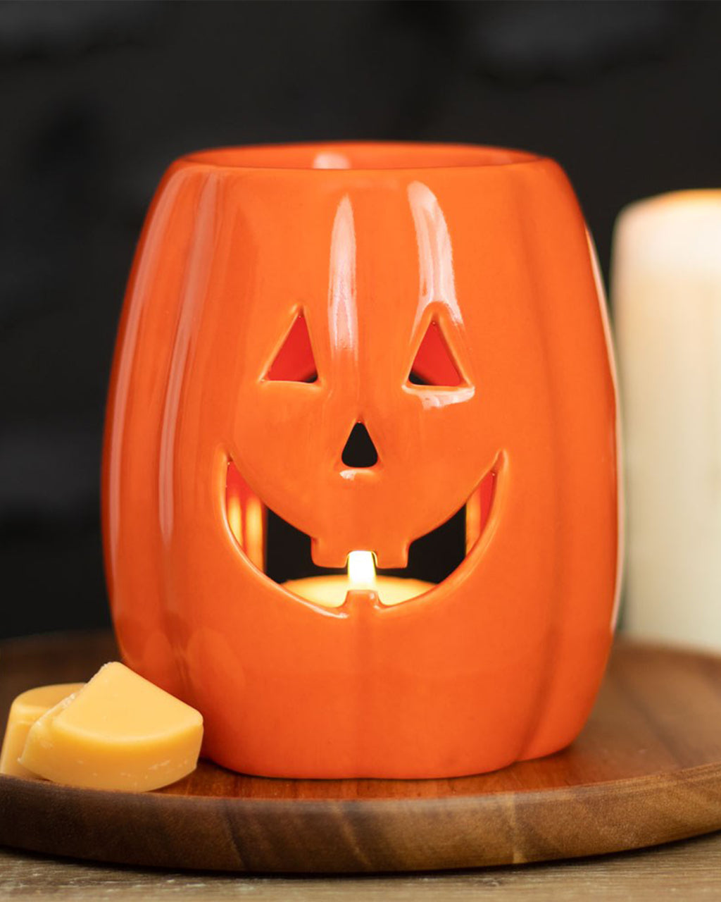 Rebel Romance ceramic Jack-o’-Lantern wax warmer glowing with a tealight, styled on a wooden board with wax melts. A festive way to enjoy autumn scents in your spooky-themed space.