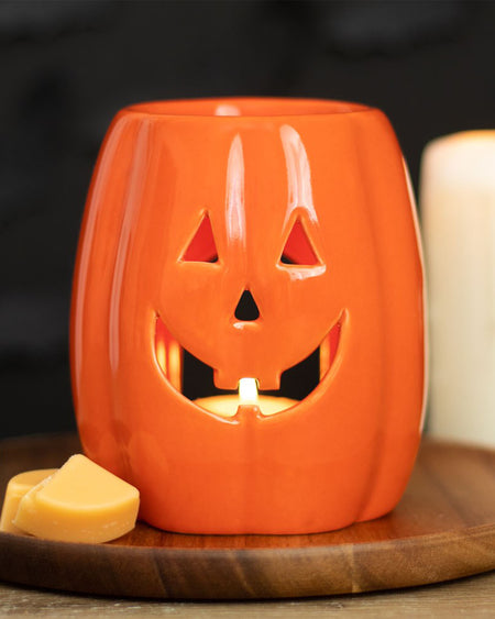 Rebel Romance ceramic Jack-o’-Lantern wax warmer glowing with a tealight, styled on a wooden board with wax melts. A festive way to enjoy autumn scents in your spooky-themed space.