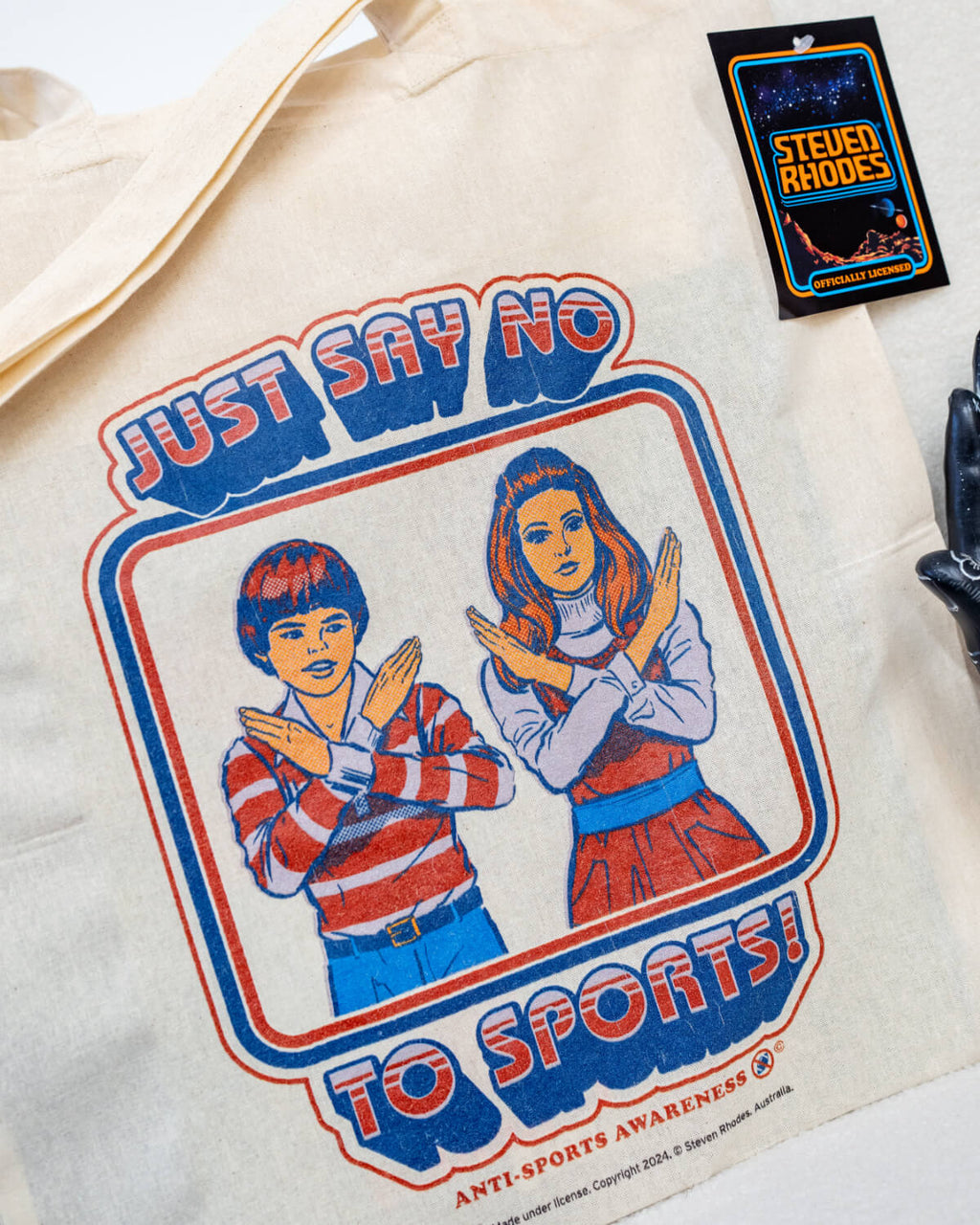 A natural cotton tote bag featuring a retro-style Steven Rhodes design with two children crossing their arms in an “X” shape and the text “Just Say No To Sports!” in bold, vintage-style lettering. The bag has a minimalist design with sturdy handles, perfect for daily use.