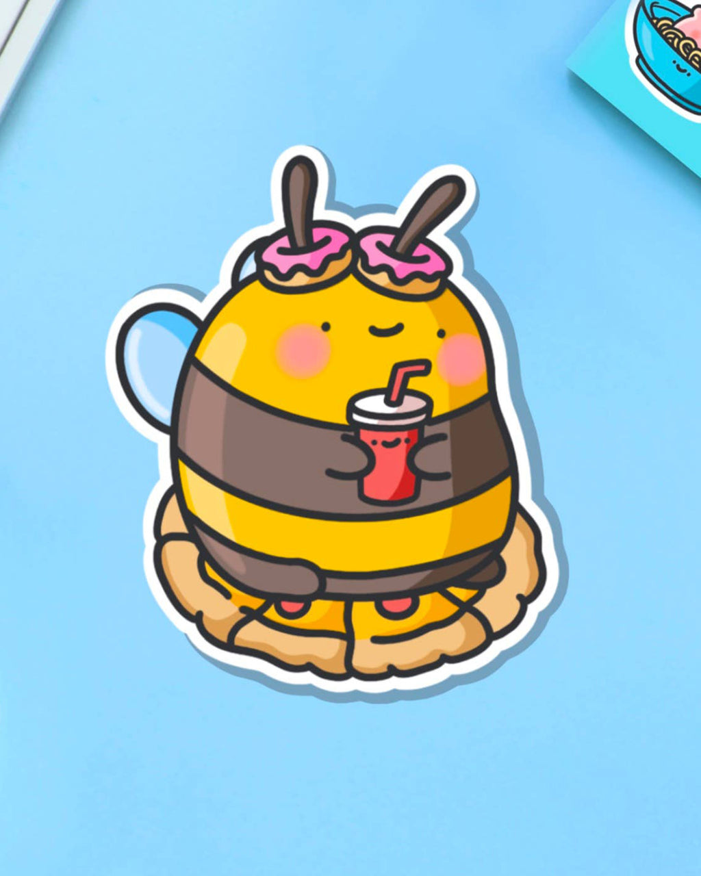 Cute cartoon-style vinyl sticker of a bumblebee with pink doughnut antennae, holding a red drink and sitting on a pie. Ideal for laptops, bottles, and journals.