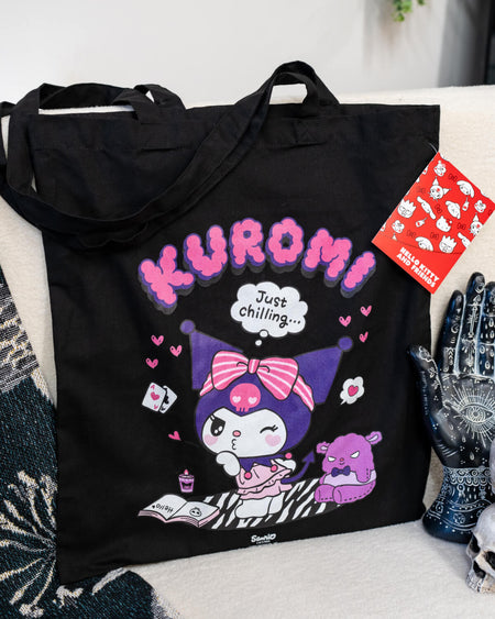 A black tote bag featuring Kuromi lounging with a zebra blanket, hearts, and a thought bubble saying “Just Chilling…” in purple and pink tones. The design captures Kuromi’s playful yet relaxed personality.