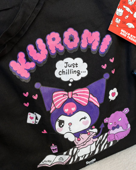 A black tote bag featuring Kuromi lounging with a zebra blanket, hearts, and a thought bubble saying “Just Chilling…” in purple and pink tones. The design captures Kuromi’s playful yet relaxed personality.