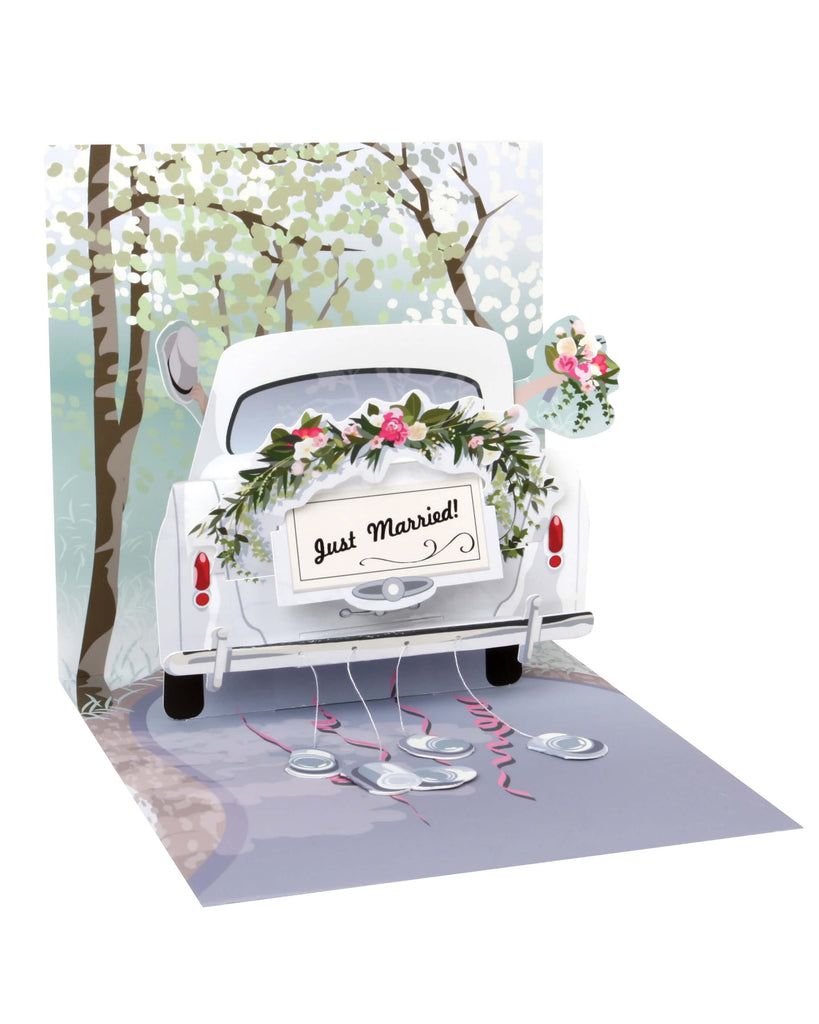 Pop-up wedding greeting card featuring a white vintage car with a “Just Married” sign, floral garland, and tin cans trailing behind on pink ribbons. The card has a spring detail for movement, glitter accents, and a scenic background of trees and a country lane. Includes a white envelope, perfect for newlyweds and wedding celebrations.