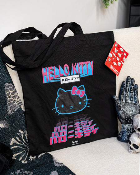 A black Hello Kitty tote bag with a Tokyo-inspired neon design in pink and blue, featuring the iconic character’s face and stylised Japanese text. The bag is laid on a cosy surface with alternative décor, including a celestial hand sculpture and small skulls. A red Hello Kitty tag is attached to the handle. Sold by Rebel Romance, a UK business celebrating alternative fashion and retro brands.