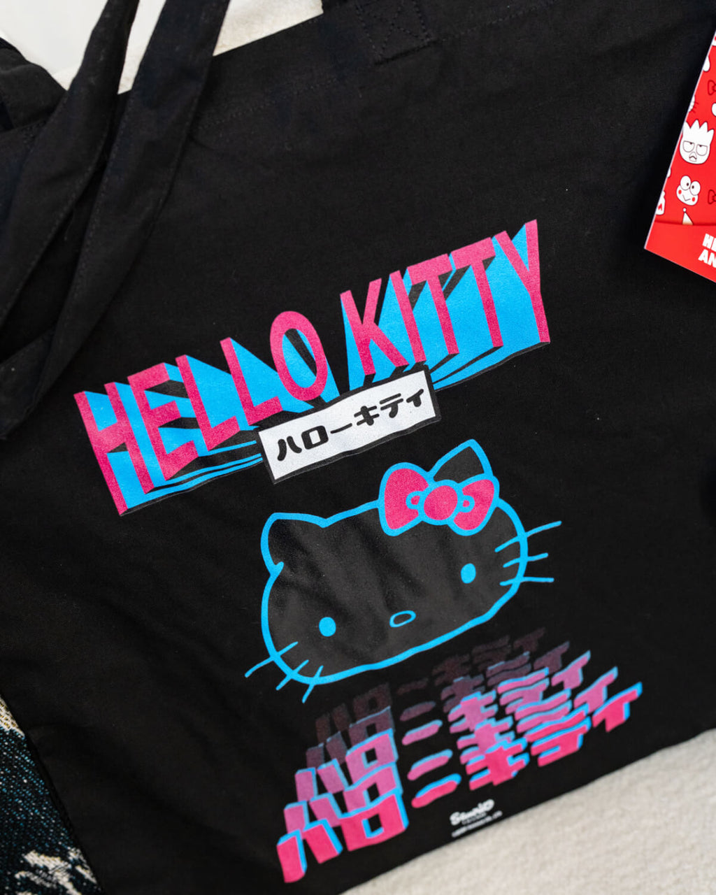 Close-up of the Hello Kitty (Tokyo) Black Tote Bag, showing its vibrant neon pink and blue design on a black fabric background. The print features a stylised Hello Kitty face, Japanese text, and bold lettering. The texture of the tote is visible, with a red Hello Kitty tag attached. Sold by Rebel Romance, a UK small business embracing alternative fashion and retro branded characters.