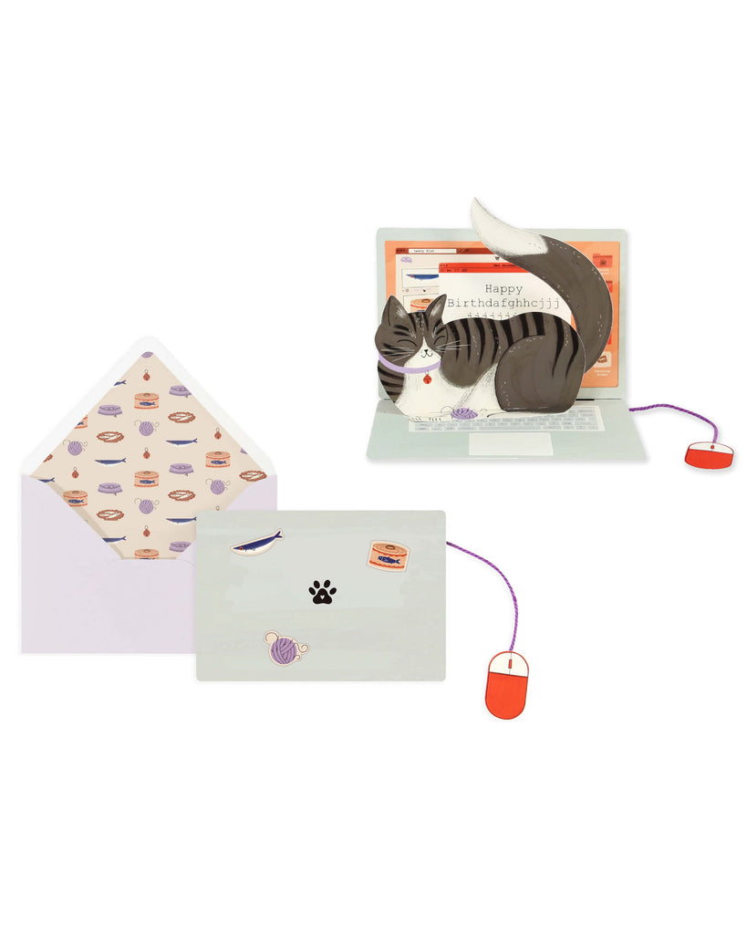 A small die-cut pop-up greeting card shaped like a laptop with a grey striped cat sitting on the keyboard. The cat’s tail curves over the screen displaying the message “Happy birthdafghhcjjjjjrrrttttt.” A red foil computer mouse is attached with a purple yarn cord, and the card comes with a lavender envelope decorated with fish, yarn, and cat food illustrations.