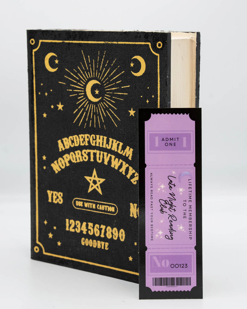 Close-up of a black and gold ouija board-style book with celestial illustrations, next to a purple ticket-shaped bookmark that reads "Lifetime Membership to the Late Night Reading Club" by Rebel Romance. A whimsical gift for alternative readers who stay up past bedtime. Designed by small UK brand Rebel Romance, known for celebrating unique and millennial-friendly aesthetics.