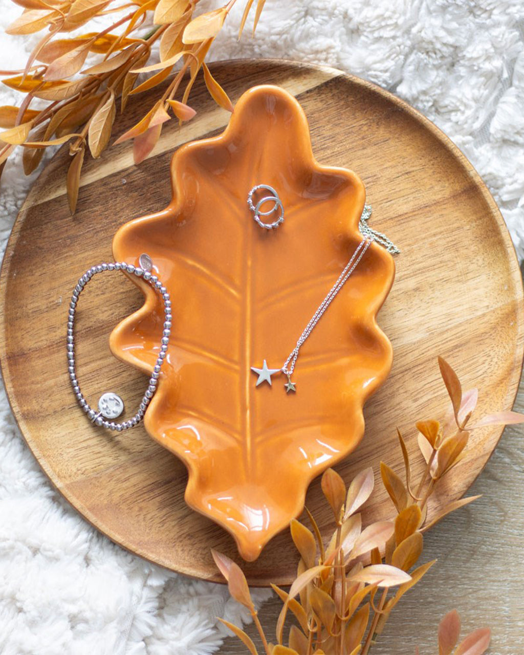 A beautifully styled shot of the ceramic autumn leaf dish, displayed on a round wooden tray. Silver jewellery pieces, including a star necklace and bracelet, rest on the dish, demonstrating its perfect use as a jewellery holder or trinket tray.