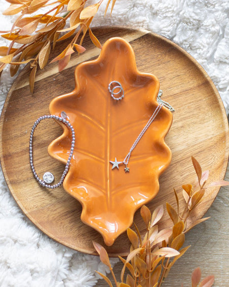A beautifully styled shot of the ceramic autumn leaf dish, displayed on a round wooden tray. Silver jewellery pieces, including a star necklace and bracelet, rest on the dish, demonstrating its perfect use as a jewellery holder or trinket tray.