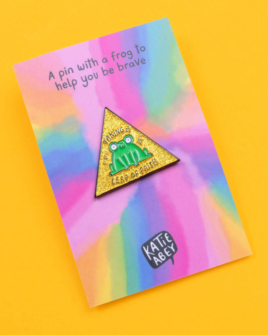 Katie Abey 'Leap of Faith' frog pin displayed on a rainbow background card. The pin features a quirky green frog on a glittery yellow triangle with the words 'Taking a Leap of Faith'. The card reads, 'A pin with a frog to help you be brave'.