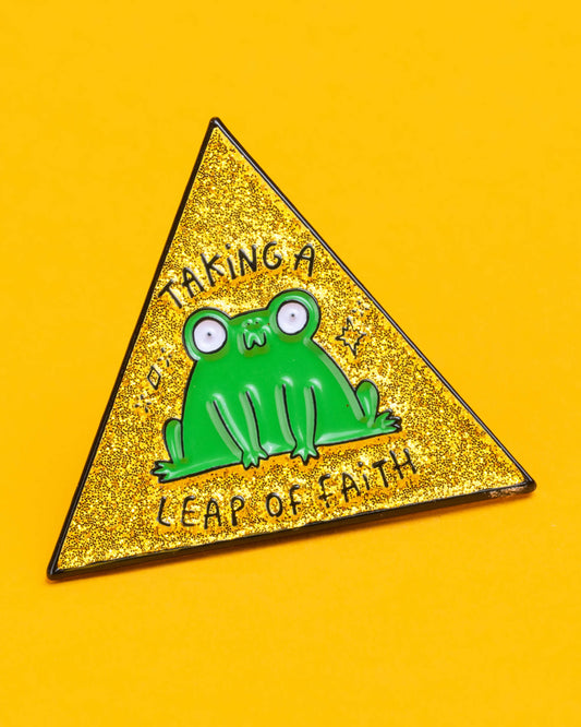Close-up of the Katie Abey 'Leap of Faith' enamel pin featuring a quirky green frog on a yellow glittery triangle background. The words 'Taking a Leap of Faith' are written around the frog.