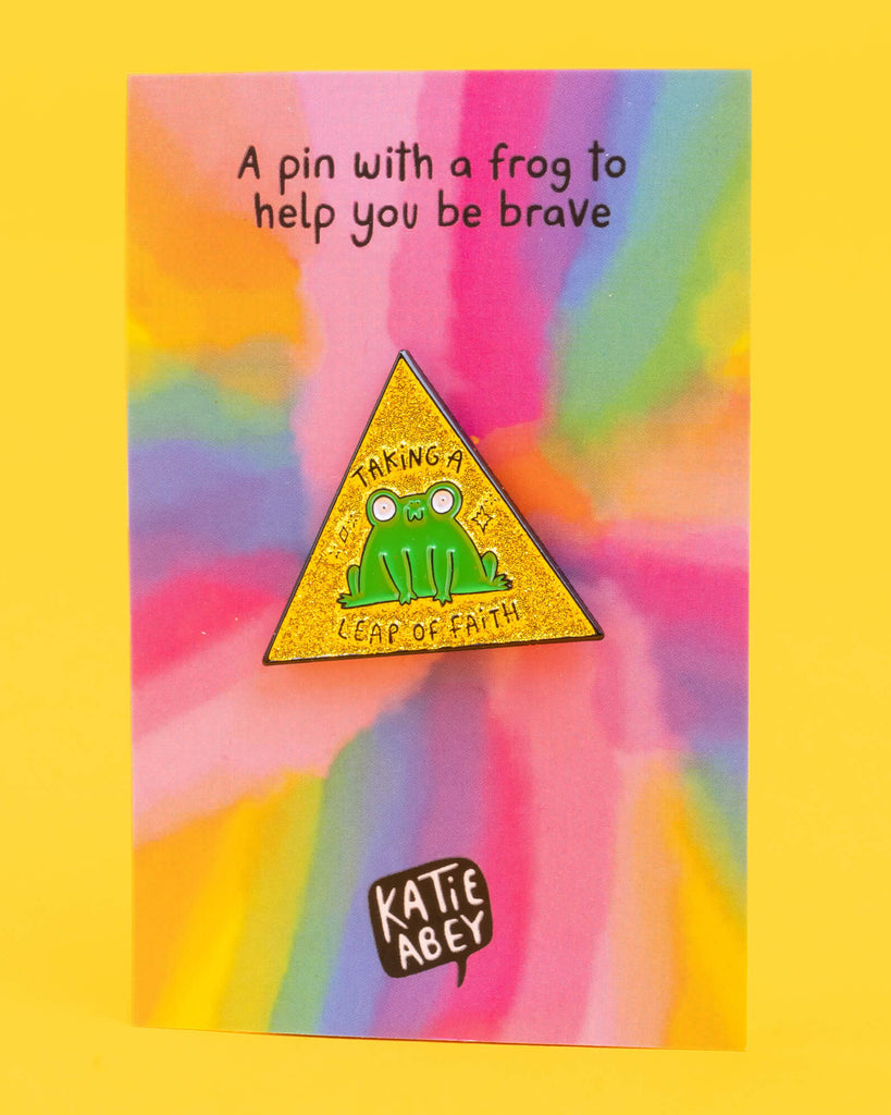 Katie Abey 'Leap of Faith' frog pin on a colourful rainbow background card. The green frog on the pin sits on a yellow glittery triangle with the words 'Taking a Leap of Faith'. Text above reads, 'A pin with a frog to help you be brave'.