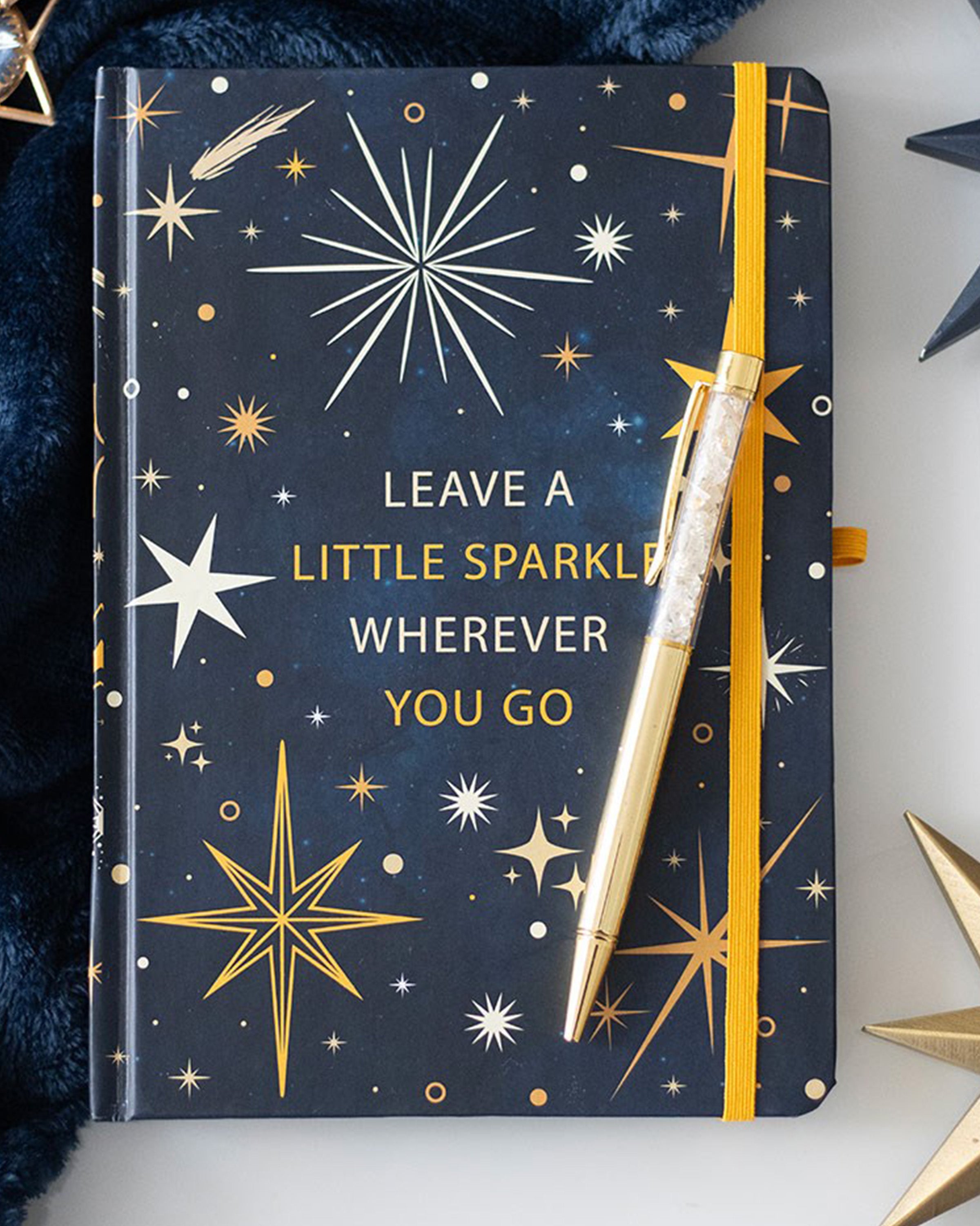 A navy blue A5 notebook with a celestial star design and the phrase “Leave a little sparkle wherever you go,” paired with a gold citrine pen, styled with gold star accessories and a navy blanket.