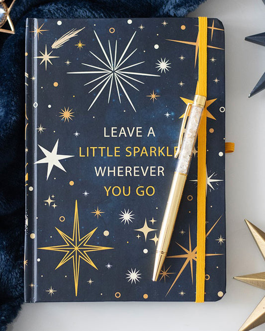A navy blue A5 notebook with a celestial star design and the phrase “Leave a little sparkle wherever you go,” paired with a gold citrine pen, styled with gold star accessories and a navy blanket.