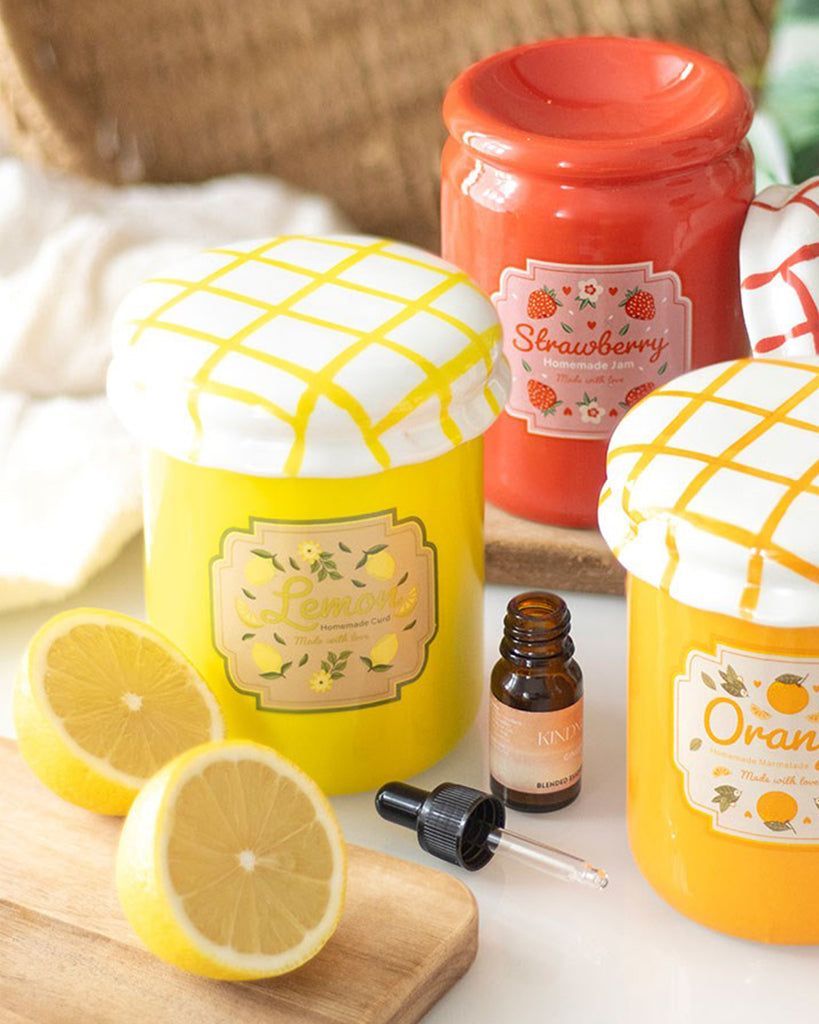 A cheerful yellow lemon curd jar-shaped oil burner and wax warmer by Rebel Romance, styled with fresh lemons, an essential oil bottle and other colourful jam jar burners in the background. A charming, vintage-inspired homeware gift for adding zest to your kitchen or living space.