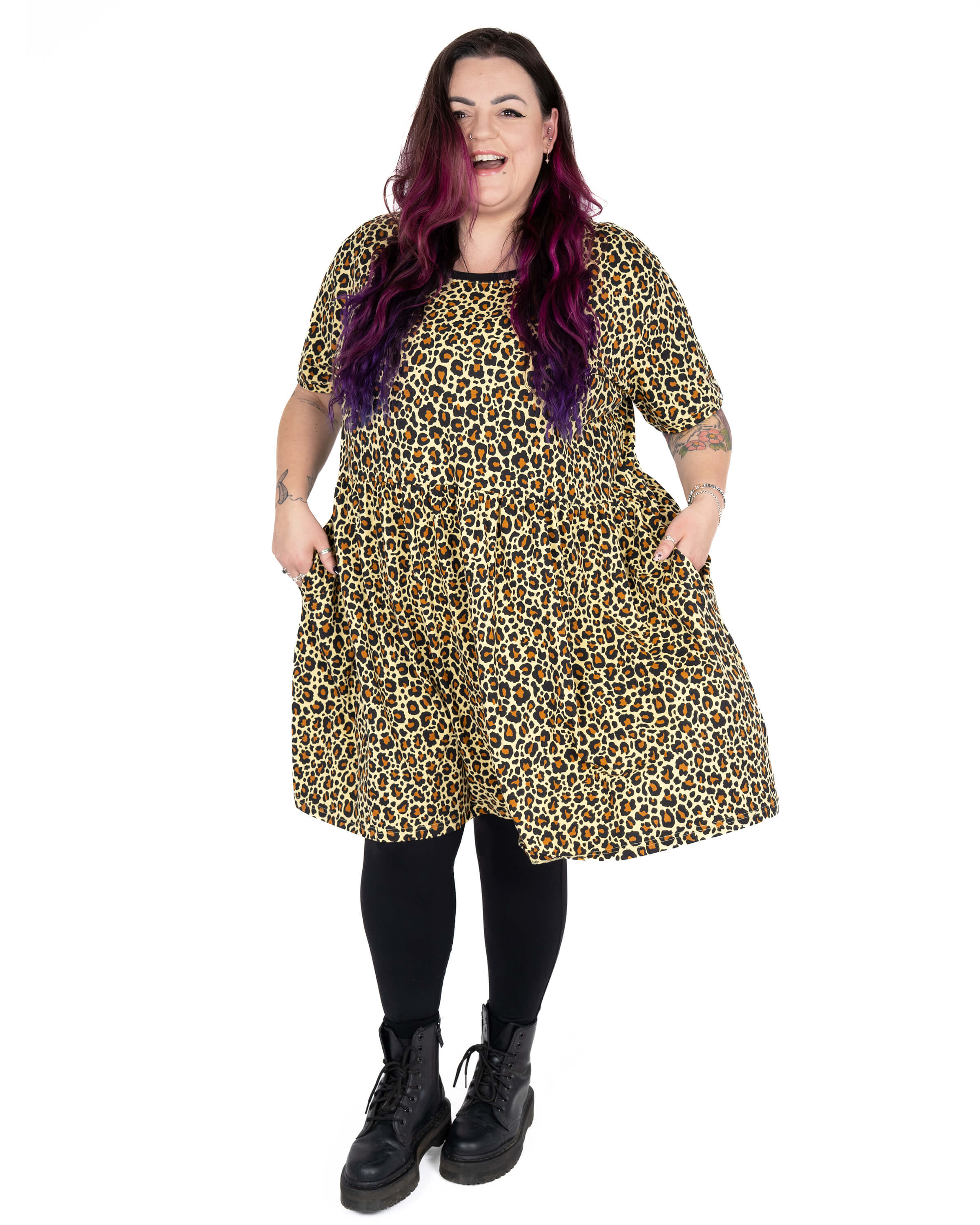 Luisa wears the Rebel Romance Classic Leopard Print Stretch Smock Dress in a front-facing pose, smiling with her hands in the pockets. This comfy short-sleeved dress features a bold all-over leopard print with a relaxed, oversized fit. Styled with black tights and chunky black boots. Luisa (she/her) is 5'6