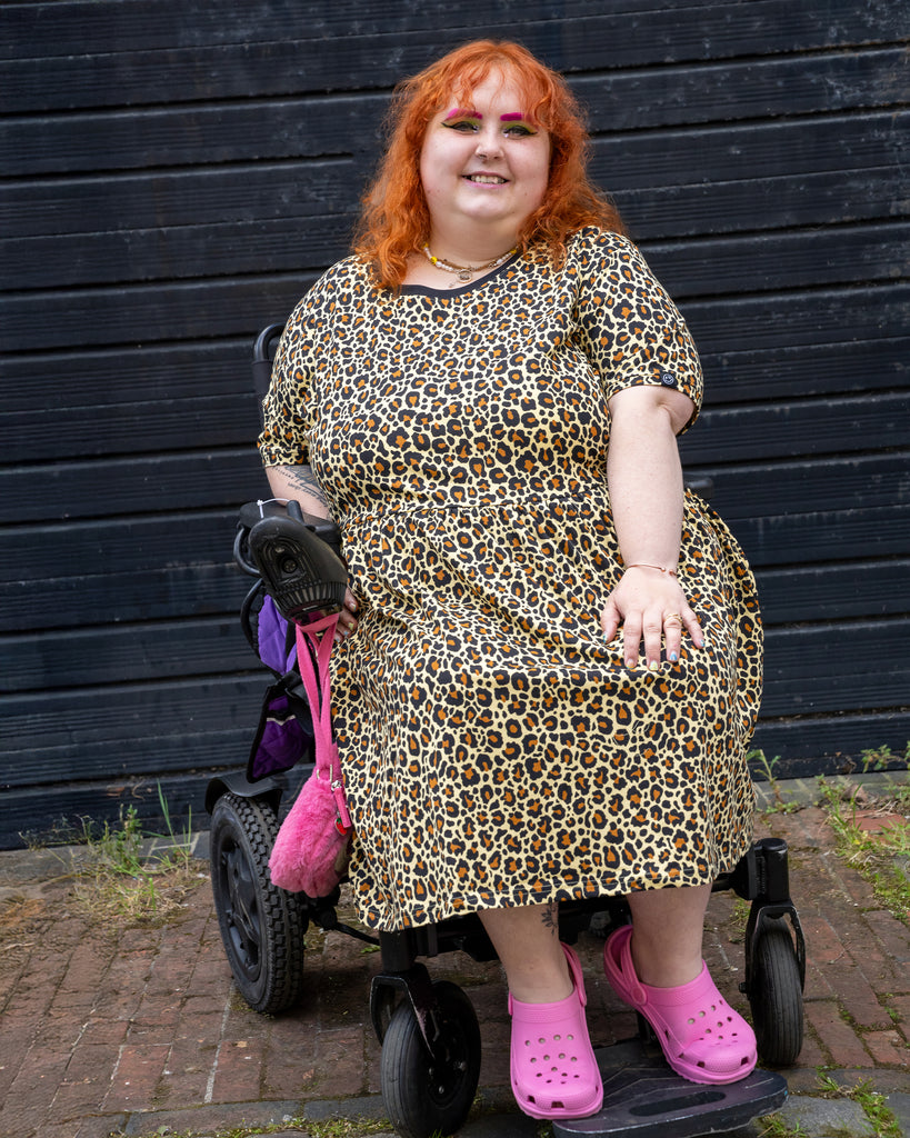 Lauren, a plus size model using a wheelchair, wearing the Rebel Romance Classic Leopard Print Stretch Smock Dress with pink Crocs and bright makeup.