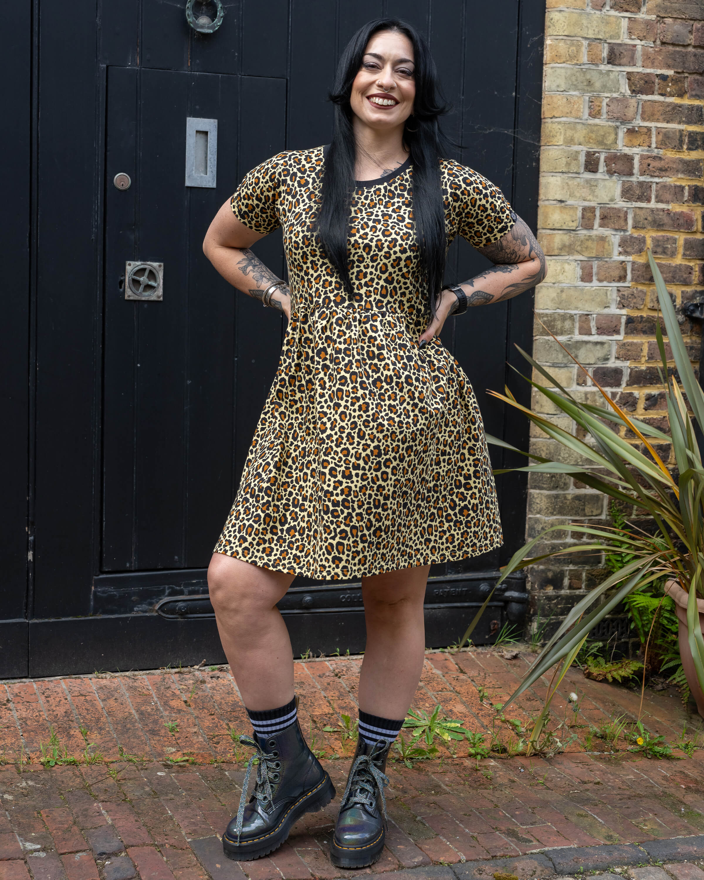 Carmen smiling and posing with their hands on their hips wearing the Rebel Romance Classic Leopard Print Stretch Smock Dress styled with chunky black boots in front of a dark wooden door.
