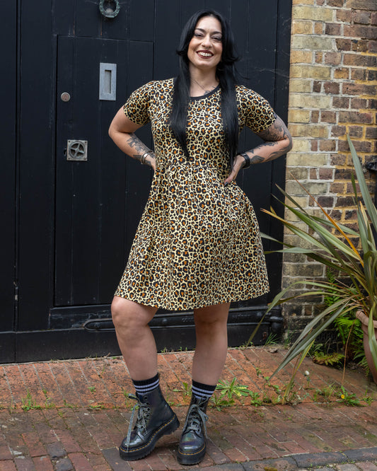 Carmen smiling and posing with their hands on their hips wearing the Rebel Romance Classic Leopard Print Stretch Smock Dress styled with chunky black boots in front of a dark wooden door.