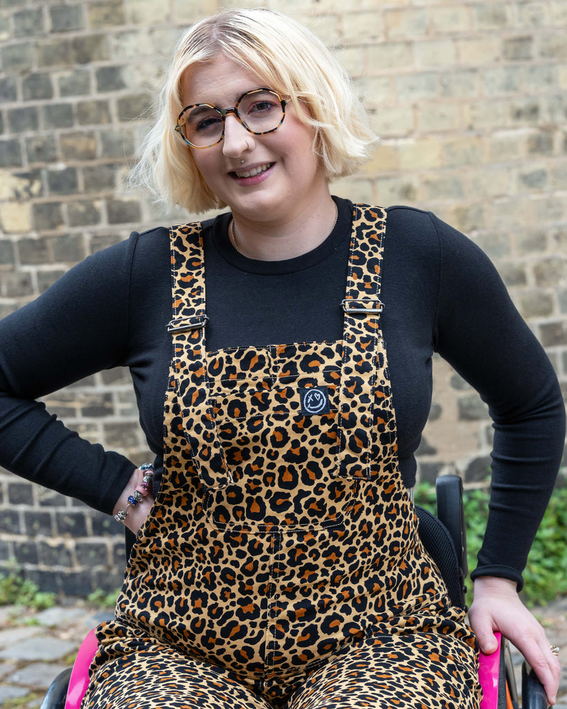 Close-up of Eliza smiling in Rebel Romance Leopard Print Dungarees, showing the chest pocket with the Rebel Romance smiley patch. Styled with a fitted black top and glasses.