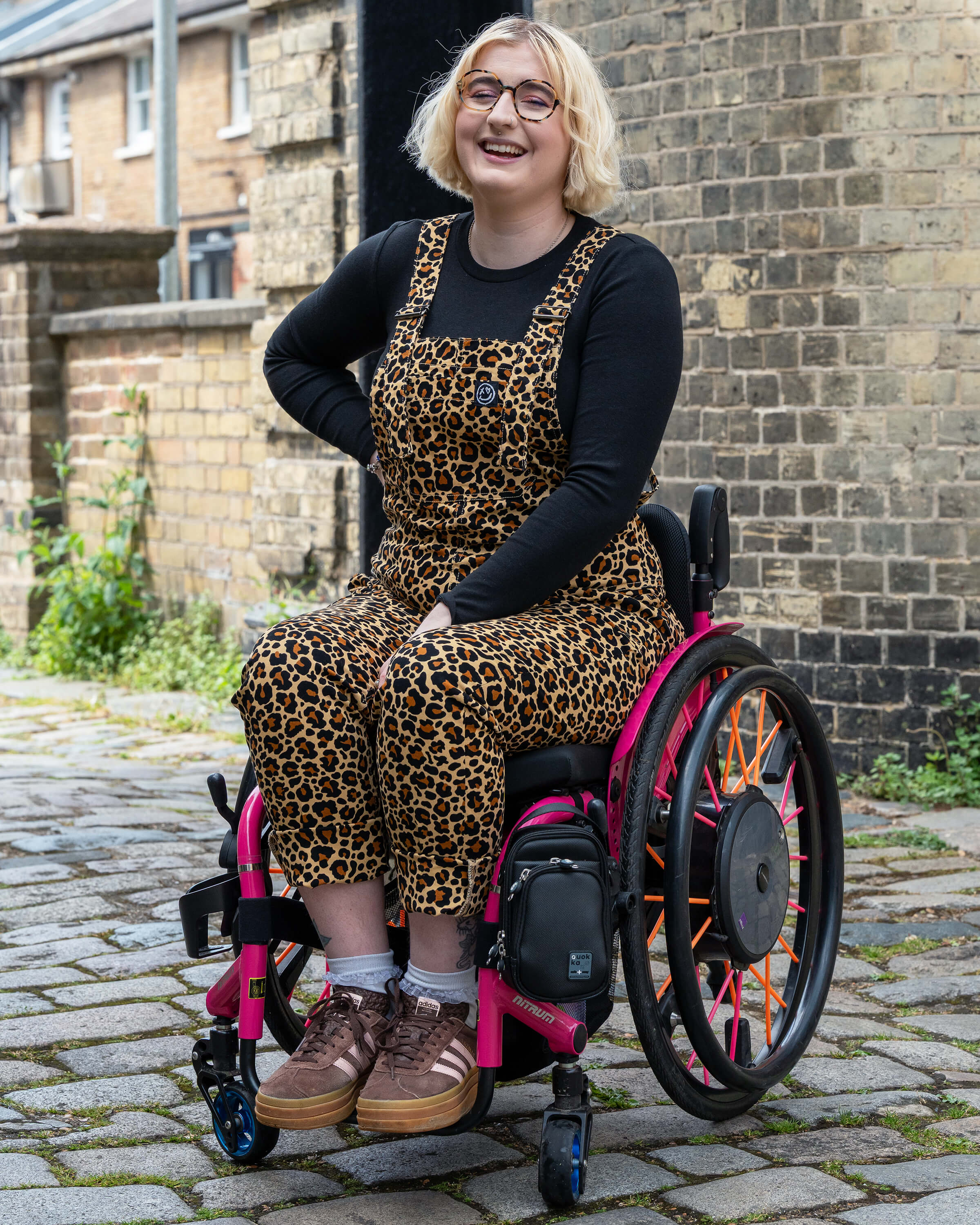 Eliza laughing wearing Rebel Romance Classic Leopard Print Dungarees in size XS. The bold print and comfy fit are styled with a black long sleeve top, sat in a vibrant pink wheelchair.