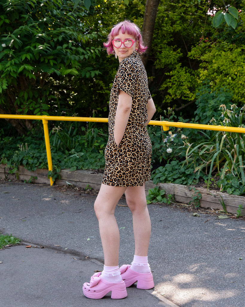 Model Jess turns slightly to show the side profile of the Rebel Romance Classic Leopard Playsuit. She smiles back at the camera with hands in her pockets, framed by leafy green plants.