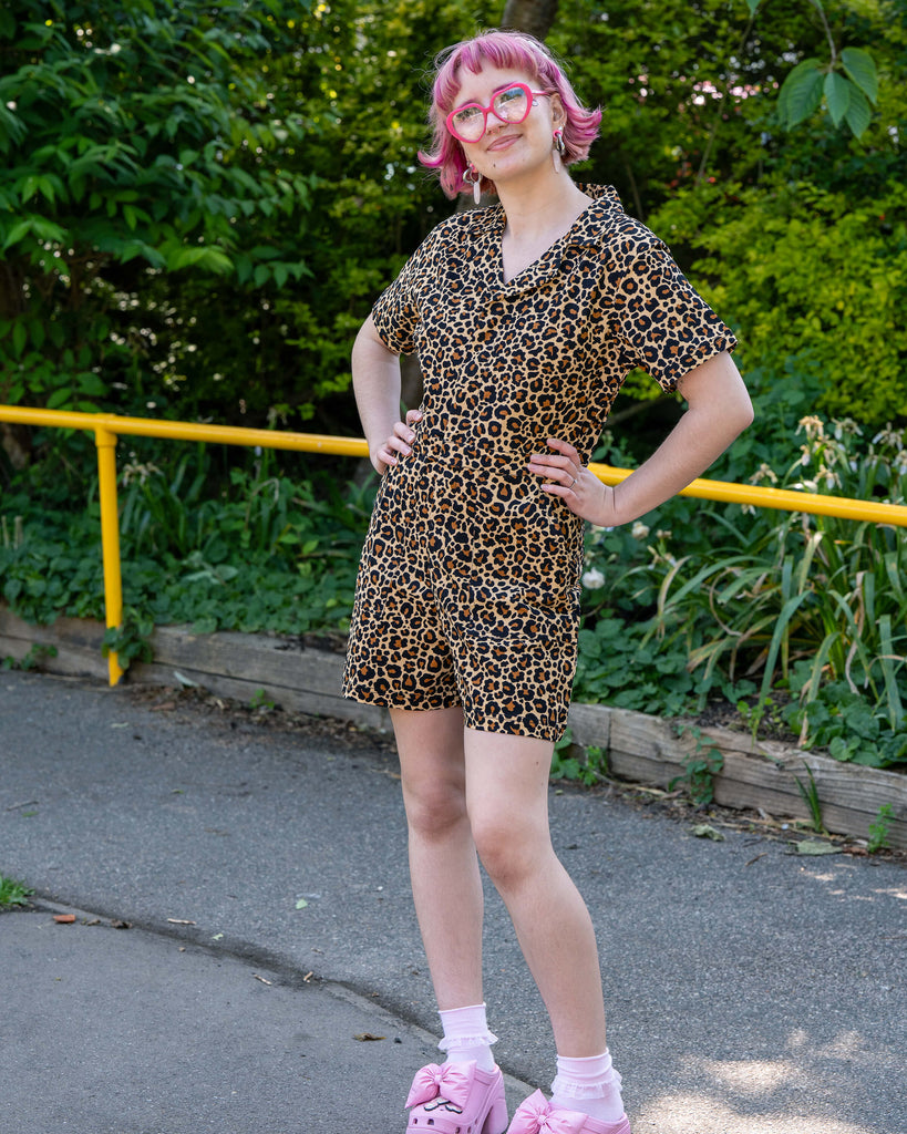 Jess poses with hands on hips in the Rebel Romance Classic Leopard Playsuit. The V-neckline, short sleeves, and structured shape are visible, paired with pink heels and a fun woodland backdrop.
