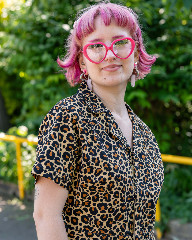 Close-up of Jess in the Rebel Romance Classic Leopard Playsuit, showing the zip-up front, collared neckline, and bold leopard print. She wears pink heart glasses and crystal earrings.