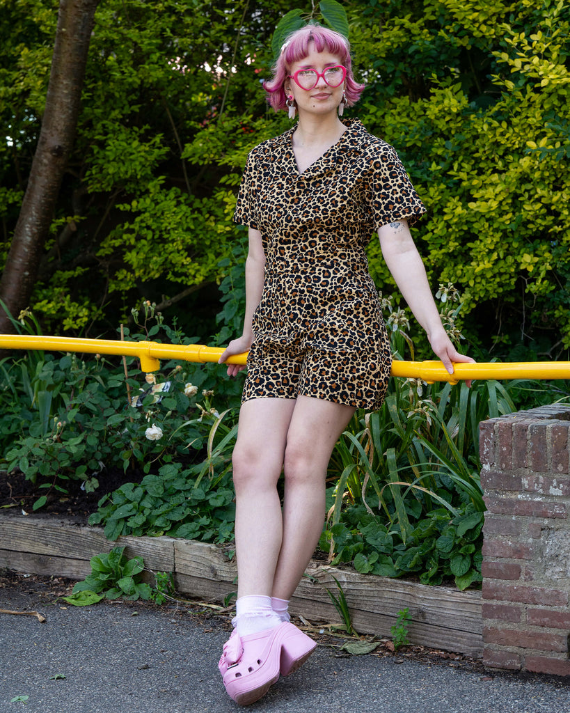 Jess stands with legs crossed and arms relaxed, modelling the Rebel Romance Classic Leopard Playsuit. She pairs it with pink chunky heels and heart-shaped glasses for a playful, alt aesthetic.
