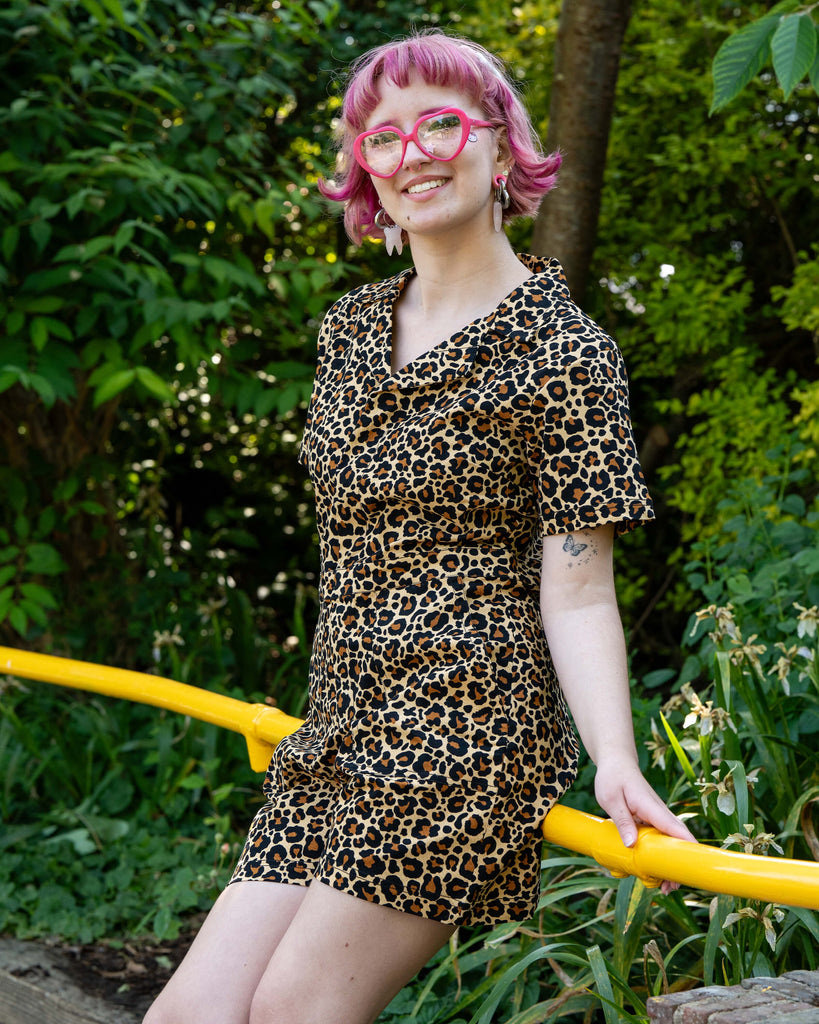 Model Jess poses in a garden setting wearing the Rebel Romance Classic Leopard Playsuit. She smiles brightly, sitting on a yellow rail with green foliage behind her, showing off the bold leopard print.