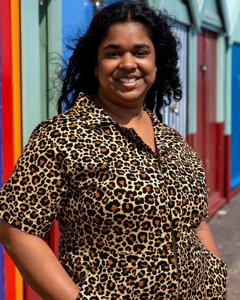 Close-up of Vidhya smiling in front of rainbow-striped beach huts while wearing the Rebel Romance Classic Leopard Playsuit. The leopard print twill fabric and structured collar are clearly shown.