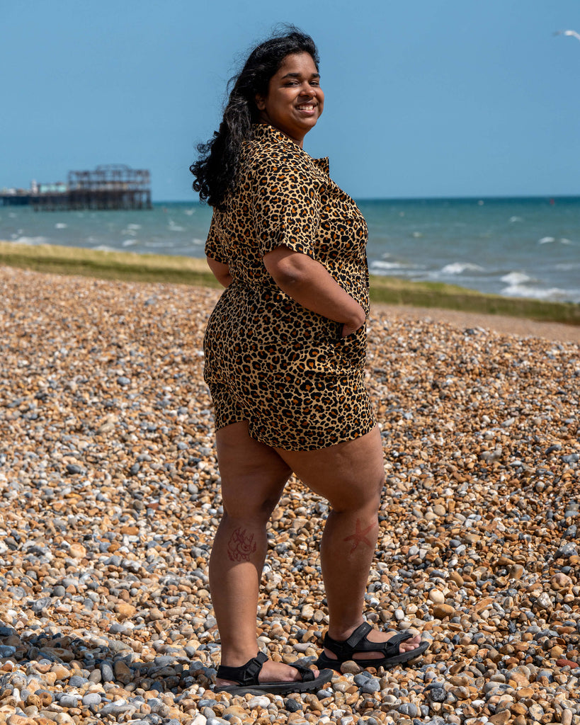 Vidhya smiles on a pebbled beach wearing the Rebel Romance Classic Leopard Playsuit. The stretchy leopard print romper has a collared neckline and side pockets. Perfect for bold alternative summer fashion.
