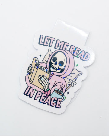 Holographic magnetic bookmark featuring a pastel skeleton in a pink hoodie reading a book with a steaming mug beside them. Surrounded by stars and the phrase “Let Me Read in Peace,” this quirky accessory is perfect for goth readers, introverts, and bookworms with a dark sense of humour. A spooky-sweet design by Rebel Romance for alternative stationery lovers.