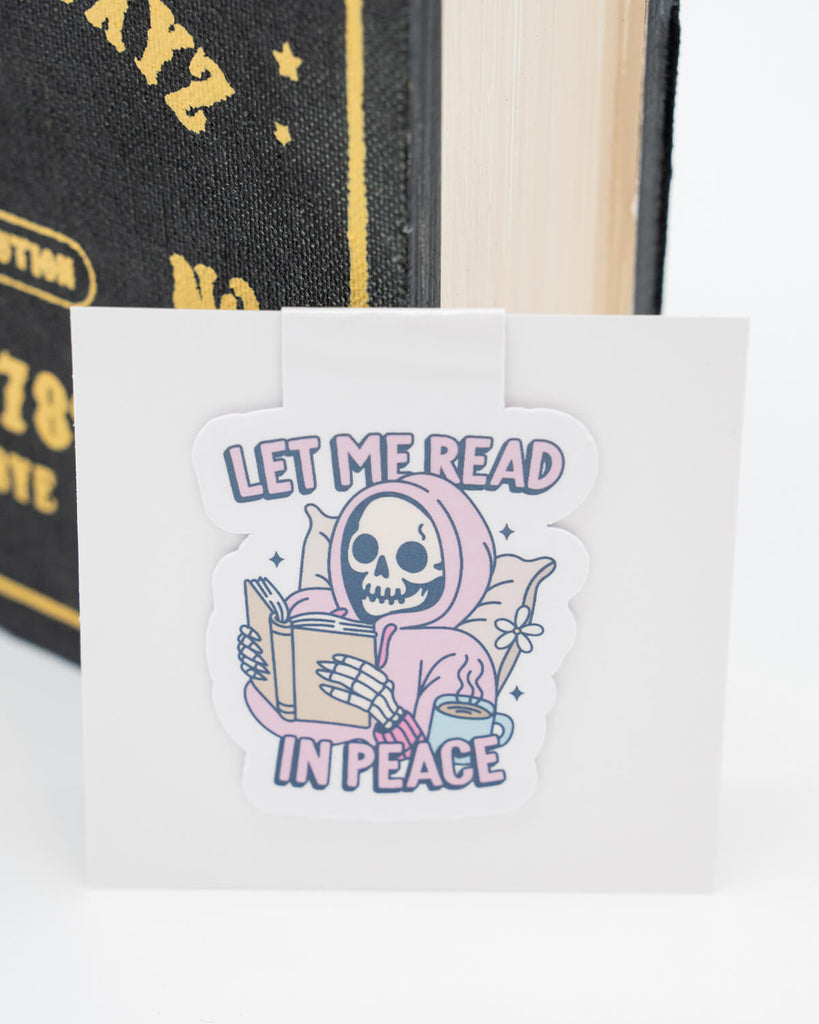 Let Me Read in Peace Magnetic Bookmark clipped to a book, showing a skeleton reading in bed with a coffee and soft pillow. Text reads “Let Me Read in Peace” in pastel tones with a holographic finish. A fun and gothic addition to any reading session, ideal for horror fans, dark academia vibes, and book lovers who prefer peace and quiet. From Rebel Romance.