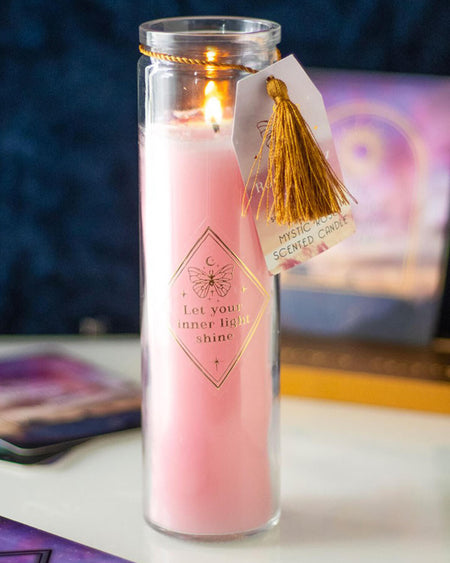 Close-up of the Mystic Rose candle lit, with a soft pink glow in a tall glass tube, decorated with gold lettering and a hanging tassel, placed on a mystical-themed surface with oracle cards in the background.