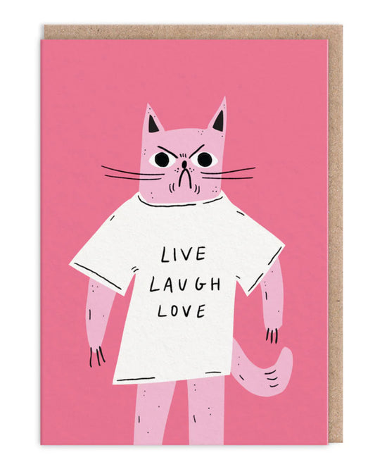 Pink illustrated greeting card from Rebel Romance featuring a grumpy pink cartoon cat in a white oversized T-shirt that reads “Live Laugh Love”. The cat stands against a solid pink background and has a hilariously angry expression, poking fun at cheesy inspirational quotes. Comes with a brown kraft envelope. Quirky and alternative card design for birthdays, just-because moments, or fellow sarcasm lovers.
