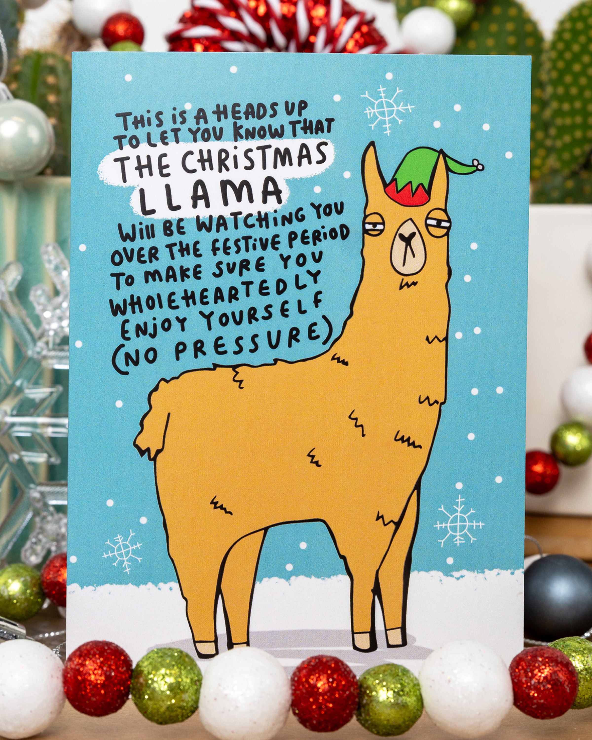 A colourful illustrated Christmas card by Rebel Romance featuring a grumpy cartoon llama in a festive elf hat, drawn by Katie Abey. Surrounded by snowflakes and baubles, the card reads: 