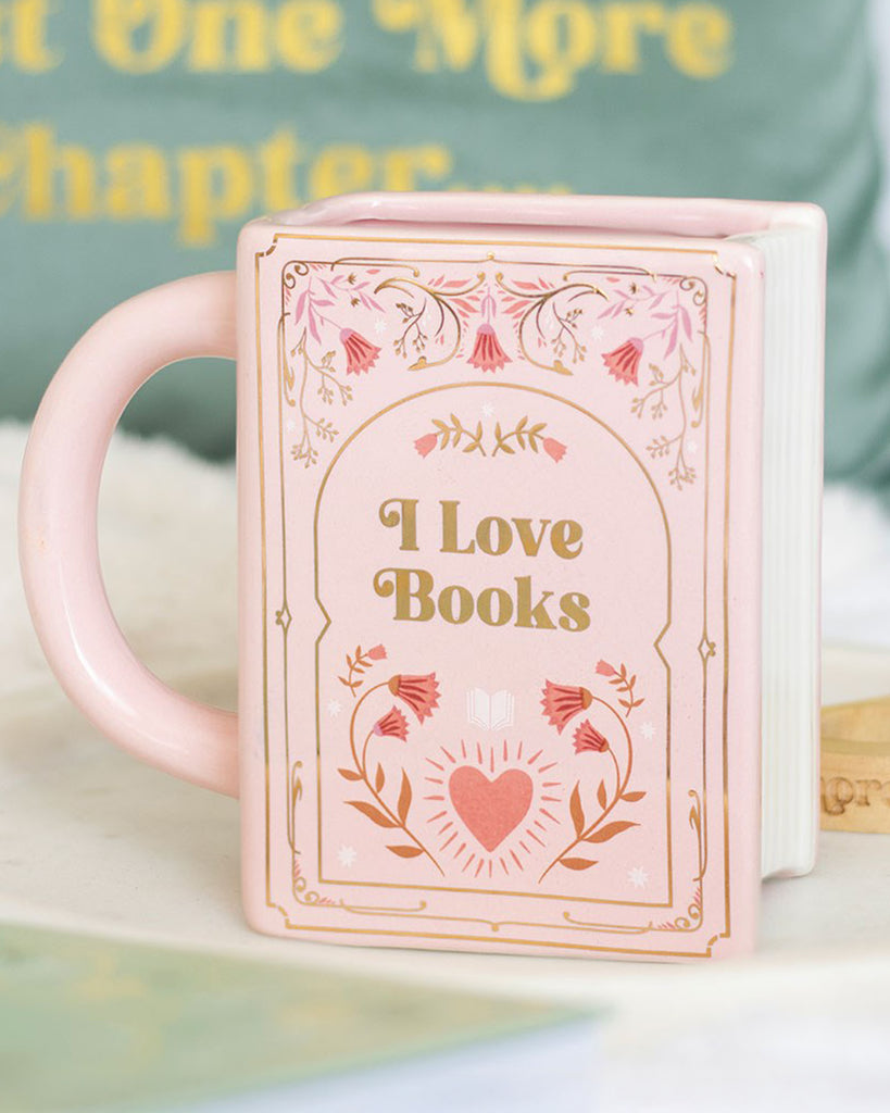 Lifestyle photo of the Rebel Romance “I Love Books” pink ceramic mug shaped like a book, styled with cosy home décor. A fun, literary-inspired gift for tea and coffee lovers.
