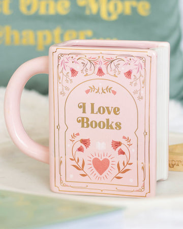 Lifestyle photo of the Rebel Romance “I Love Books” pink ceramic mug shaped like a book, styled with cosy home décor. A fun, literary-inspired gift for tea and coffee lovers.