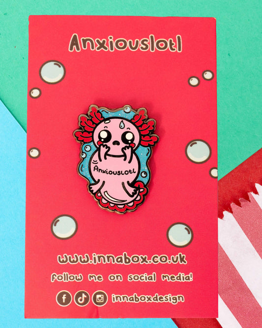 The Anxiouslotl Glittery Axolotl Enamel Pin is displayed on a red backing card that says "Anxiouslotl" with bubble graphics. This enamel pin from Rebel Romance, designed by Innabox, features a nervous-looking pink axolotl on a glittery blue background—perfect for mental health awareness and pin collectors.