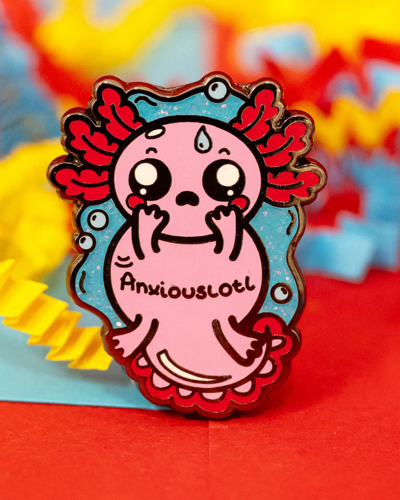 A close-up of the Anxiouslotl Glittery Axolotl Enamel Pin from Rebel Romance, designed by Innabox. The pin features a cute pink axolotl with anxious eyes and red gills, set against a glittery blue background. Surrounded by colourful shredded paper in red, yellow and blue. A quirky gift that raises awareness for anxiety and invisible struggles.
