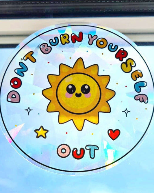 Close-up of a circular holographic window sticker from Rebel Romance featuring a cute smiling sun and the phrase “Don’t Burn Yourself Out” in colourful bubble letters. The sticker is placed on a sunlit window and casts a gentle rainbow glow. Designed by Innabox.
