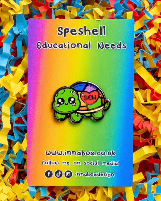 Close-up of the Speshell Educational Needs tortoise enamel pin on a rainbow gradient backing card with colourful shredded paper in the background. The pin, designed by Innabox , features a cute green tortoise with big sparkly eyes and a rainbow shell marked with a red heart and the letters "SEN". Designed to raise awareness for those with additional needs 