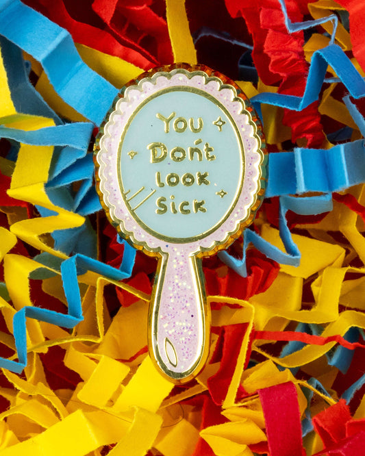 A close-up of a pastel glitter enamel pin shaped like a mirror with the phrase "You Don’t Look Sick" in gold text. Surrounded by colourful red, yellow, and blue shredded paper. 