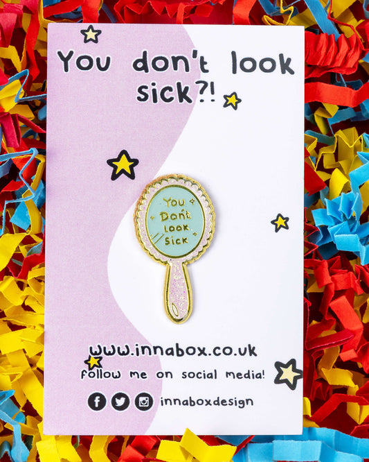 A glittery enamel pin shaped like a pink and blue hand mirror with gold edges, displayed on a backing card that reads "You don’t look sick?!". The mirror pin says "You Don’t Look Sick" in gold lettering. Designed by Innabox and sold by Rebel Romance, a UK brand celebrating alternative fashion, mental health awareness, and invisible disabilities.