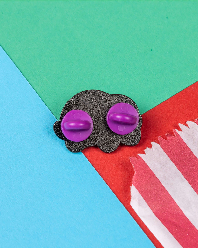 Back view of the Speshell Educational Needs enamel pin by Innabox. The pin is placed on colourful paper and features two purple rubber backs for secure fastening. Available from Rebel Romance, a UK-based brand selling alternative fashion and accessories that celebrate individuality and neurodiversity.