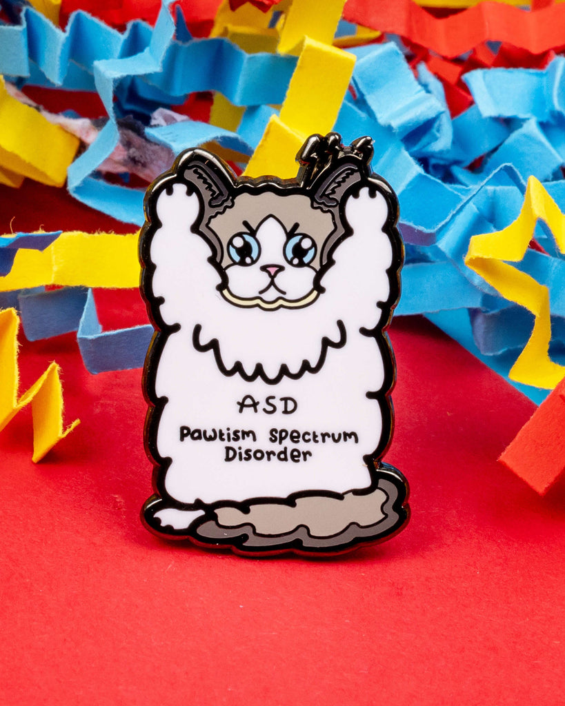 Cute cat-themed enamel pin on red background with colourful confetti strips. The pin features a fluffy cat with blue eyes, paws raised, and the words "ASD Pawtism Spectrum Disorder". Sold by Rebel Romance, a UK small business celebrating alternative fashion and raising awareness for autism in a playful, kind way.
