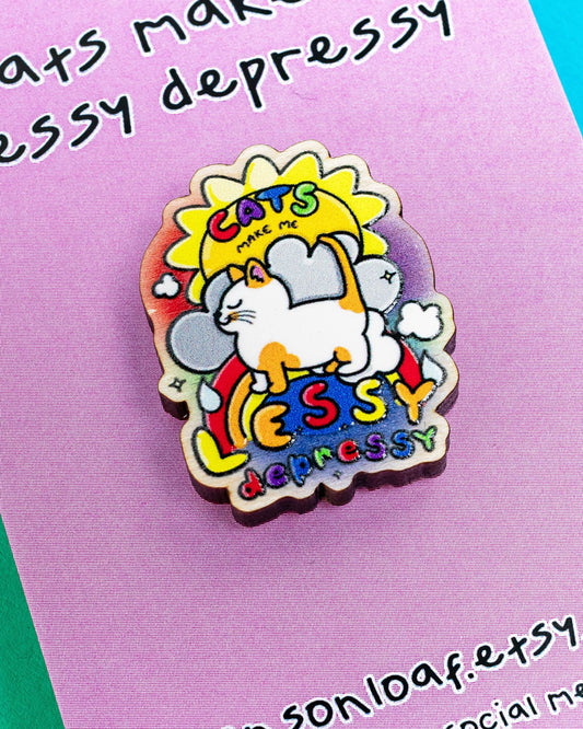 Close-up of a colourful wooden pin badge on a lilac backing card reading "Cats make me lessy depressy." The pin features a white and ginger cat standing on a rainbow with a happy sun behind it. From Rebel Romance, a small UK brand celebrating alternative fashion and gifts.