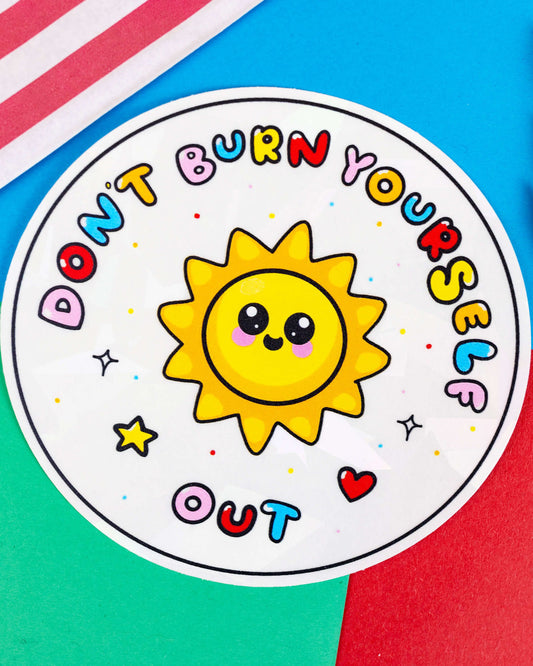 Flat lay of the Rebel Romance “Don’t Burn Yourself Out” holographic window sticker by Innabox, on a bright background. The sticker features a cheerful yellow sun and multicoloured lettering with a cracked ice effect.
