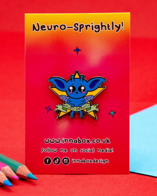 A Neuro-Sprightly enamel pin by Innabox lying on a red surface beside colourful pencils. The pin features a blue creature with wings, blushing cheeks, and a yellow star behind it, with a banner reading “Neuro-Sprightly.” Sold by Rebel Romance, a UK alternative fashion and lifestyle brand.